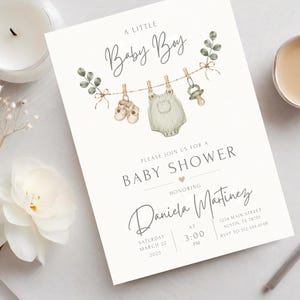 Puede incluir: Una tarjeta de invitaci&oacute;n blanca para baby shower con ilustraciones en acuarela de zapatos de beb&eacute;, un mameluco y un chupete colgados de una cuerda con hojas de eucalipto. El texto dice A LITTLE Baby Boy y PLEASE JOIN US FOR A BABY SHOWER.