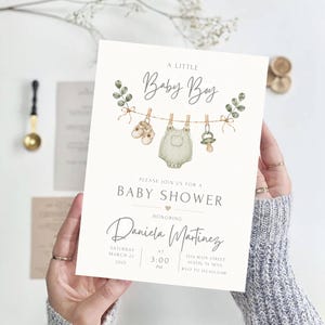 Puede incluir: Una tarjeta de invitaci&oacute;n para baby shower de color blanco con ilustraciones en acuarela de zapatitos, un mameluco y un chupete colgados de un tendedero con ramas de eucalipto. El texto dice A LITTLE Baby Boy, BABY SHOWER y DANIELA MARTINEZ.