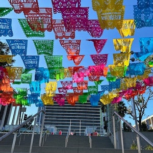 May include: A vibrant display of colorful papel picado banners strung overhead. The banners, in shades of red, pink, yellow, green, and blue, are intricately cut with decorative patterns. The text "Gloria Molina Grand Park" is visible on some banners.