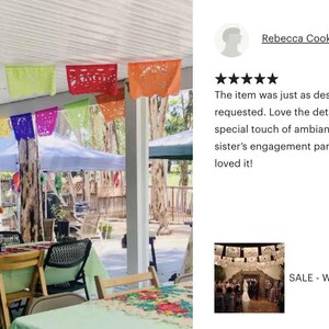 May include: A festive outdoor scene with colorful papel picado banners strung across a covered patio. Tables are set with green tablecloths, and chairs are arranged for a gathering. The scene suggests a celebration or party.