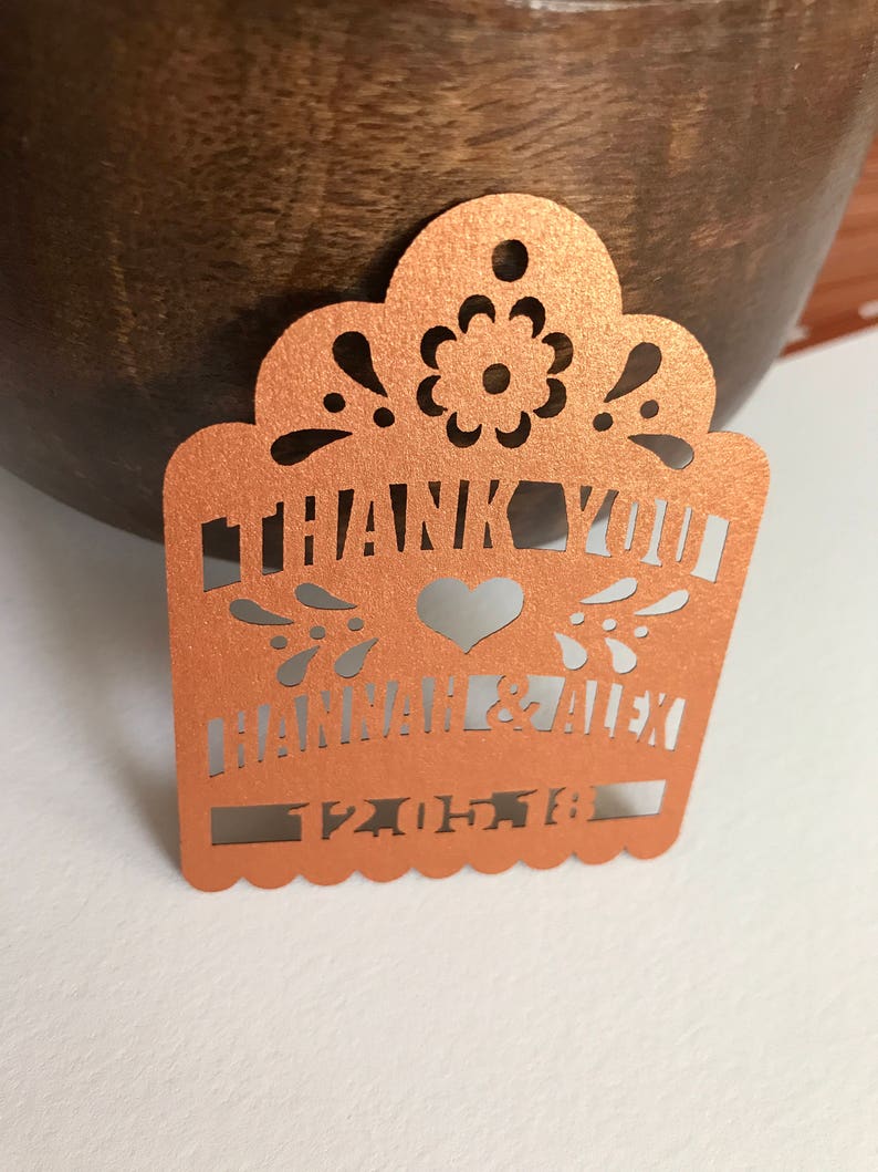 Puede incluir: Etiqueta de corte l&aacute;ser color cobre con las palabras "THANK YOU" y los nombres "HANNAH & ALEX" junto con una fecha. La etiqueta presenta un dise&ntilde;o floral y de coraz&oacute;n decorativo, ideal para recuerdos de boda.