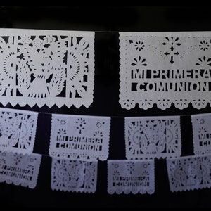 May include: White paper cutouts with intricate designs, including flowers and a cross, strung together to form a banner. The cutouts have the text "MI PRIMERA COMUNION" written on them.