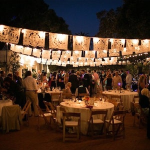 May include: Outdoor evening event with round tables set for dining. Guests mingle under strings of illuminated, decorative paper banners. The scene is lit by overhead lights and candles, creating a festive atmosphere. The event appears to be a celebration or party.