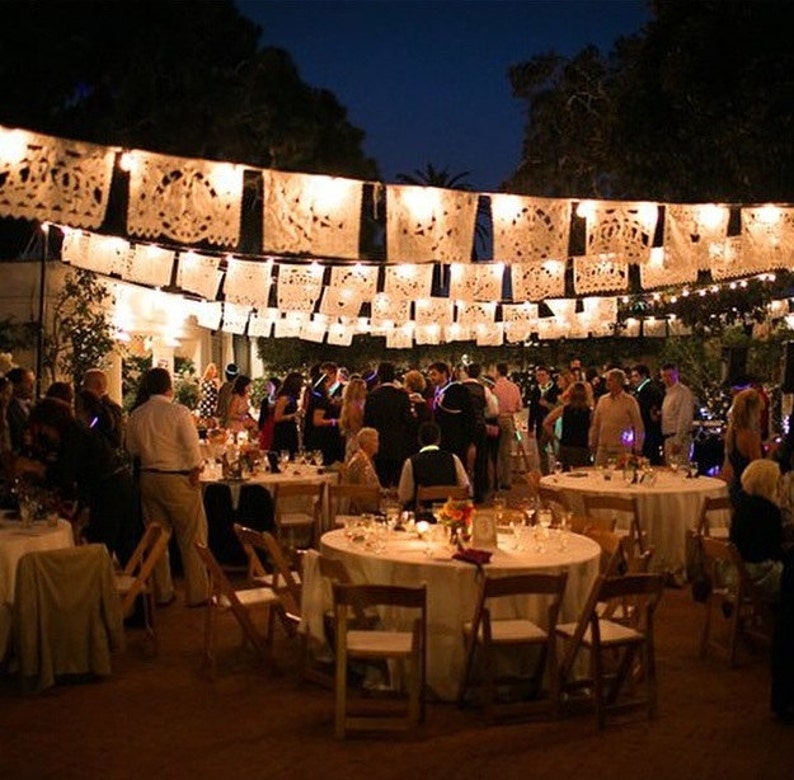 May include: Outdoor evening event with round tables set for dining. Guests mingle under strings of illuminated, decorative paper banners. The scene is lit by overhead lights and candles, creating a festive atmosphere. The event appears to be a celebration or party.