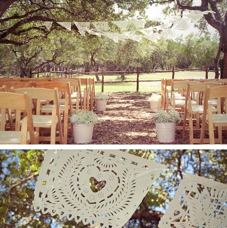 May include: An outdoor wedding ceremony setup with wooden chairs arranged on either side of a pathway. White paper banners hang overhead, and white flowers in metal buckets line the aisle. The scene is set in a wooded area.