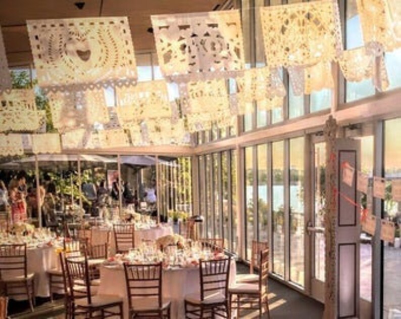 May include: An event space decorated with cream-colored papel picado banners. The banners are hung from the ceiling and feature intricate cut-out designs. Tables are set with white tablecloths, glassware, and centerpieces. Large windows offer a view of the outdoors.
