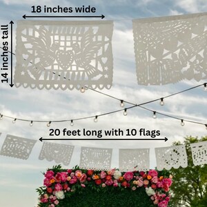 May include: White paper banner with intricate cut-out designs, 18 inches wide and 14 inches tall. The banner is 20 feet long with 10 flags. A string of lights hangs above the banner, with a floral arrangement below.