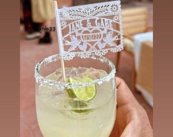 May include: A margarita cocktail in a glass with a salt-rimmed edge, a lime wedge, and a decorative white paper flag. The flag reads "JANE & GABE" and "6/10/22". The drink is held by a person.