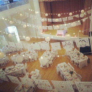 May include: An overhead view of a decorated event space. White tables and chairs are arranged for a gathering, adorned with white paper cutouts and string lights. Large paper lanterns hang from the ceiling, creating a festive atmosphere.