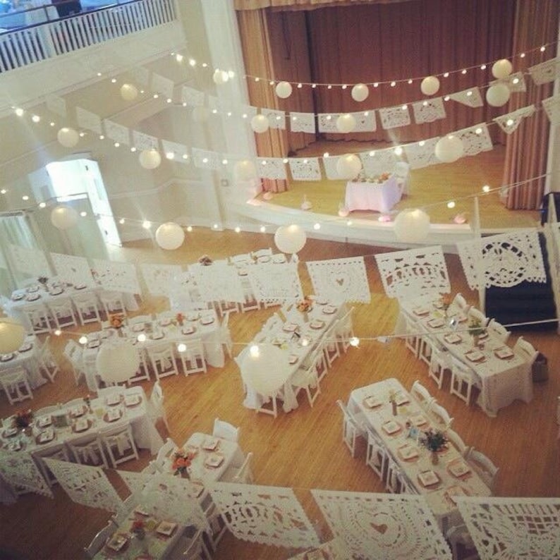 May include: An overhead view of a decorated event space. White tables and chairs are arranged for a gathering, adorned with white paper cutouts and string lights. Large paper lanterns hang from the ceiling, creating a festive atmosphere.