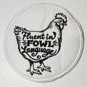 May include: White embroidered patch with a black line drawing of a chicken. The text "Fluent in Fowl Language" is written on the chicken.