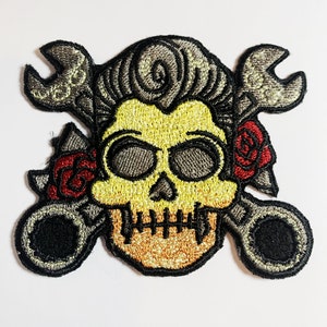 May include: Embroidered patch featuring a yellow skull with black eye sockets and a detailed jawline. Two wrenches cross behind the skull, with red roses on either side. The patch has a black border.