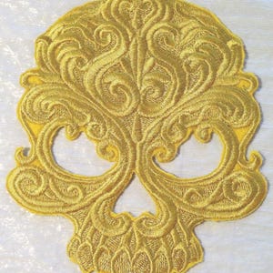 May include: A yellow embroidered skull patch with intricate floral designs. The skull is outlined in a thick yellow thread and the details are filled in with a lighter yellow thread.