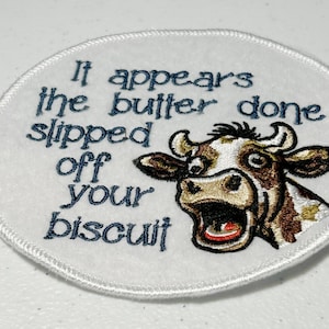 May include: White embroidered patch with a cow's face and the text "It appears the butter done slipped off your biscuit".