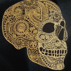 May include: Gold embroidered skull with intricate paisley and floral patterns on a black background.