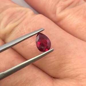 May include: A deep red, pear-shaped gemstone is held by silver tweezers. The gem is faceted, catching the light. The background is a blurred view of skin. The gem's cut reveals multiple facets.