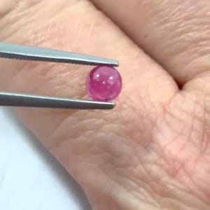 May include: A small, round, polished ruby gemstone held by tweezers. The ruby is a deep pinkish-red colour and appears translucent. The tweezers are silver-coloured and are holding the gemstone against a blurred background.