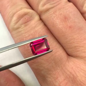 Könnte beinhalten: Ein leuchtend roter, rechteckig geschliffener Rubin wird von einer silbernen Pinzette gehalten. Der Edelstein hat ein klares, transparentes Aussehen und zeigt seine Facetten. Die Pinzette wird von einer Hand gehalten.
