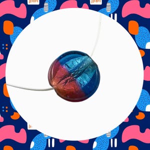 May include: A round pendant necklace with a colour gradient of blue, red, and blue. The pendant is attached to a thin white cord. The pendant has a textured surface against a white background.