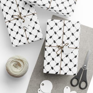 Könnte beinhalten: Drei weiße Geschenkboxen, verpackt in schwarz-weiß gemustertem Papier mit braunem Garn und weißen Geschenkanhängern. Die Boxen befinden sich auf einer grauen Oberfläche mit weißen Geschenkanhängern und einer schwarzen Schere.