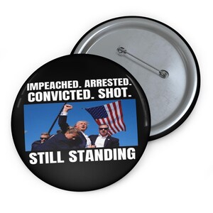 May include: A black and white button with a photo of a man in a suit holding a flag. The text on the button reads "Impeached. Arrested. Convicted. Shot. Still Standing."