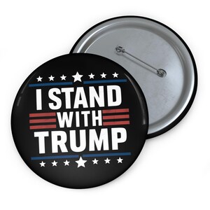 May include: A black button with a white border and stars. The button says "I Stand With Trump" in white letters with red and blue stripes.