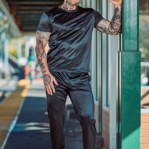 May include: A black satin t-shirt and matching pants set. The short-sleeved top and pants have a sheen. The person is wearing black and gold sneakers. The person has tattoos on their arms, neck, and hands.
