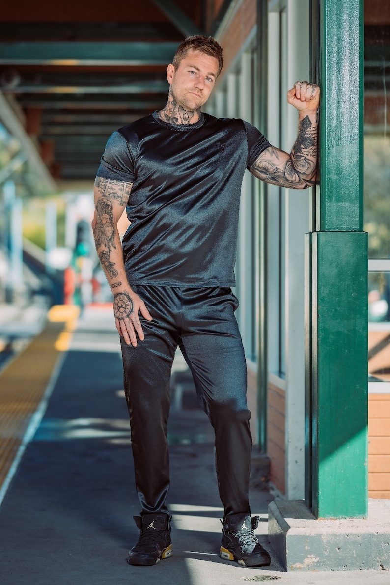 May include: A black satin t-shirt and matching pants set. The short-sleeved top and pants have a sheen. The person is wearing black and gold sneakers. The person has tattoos on their arms, neck, and hands.