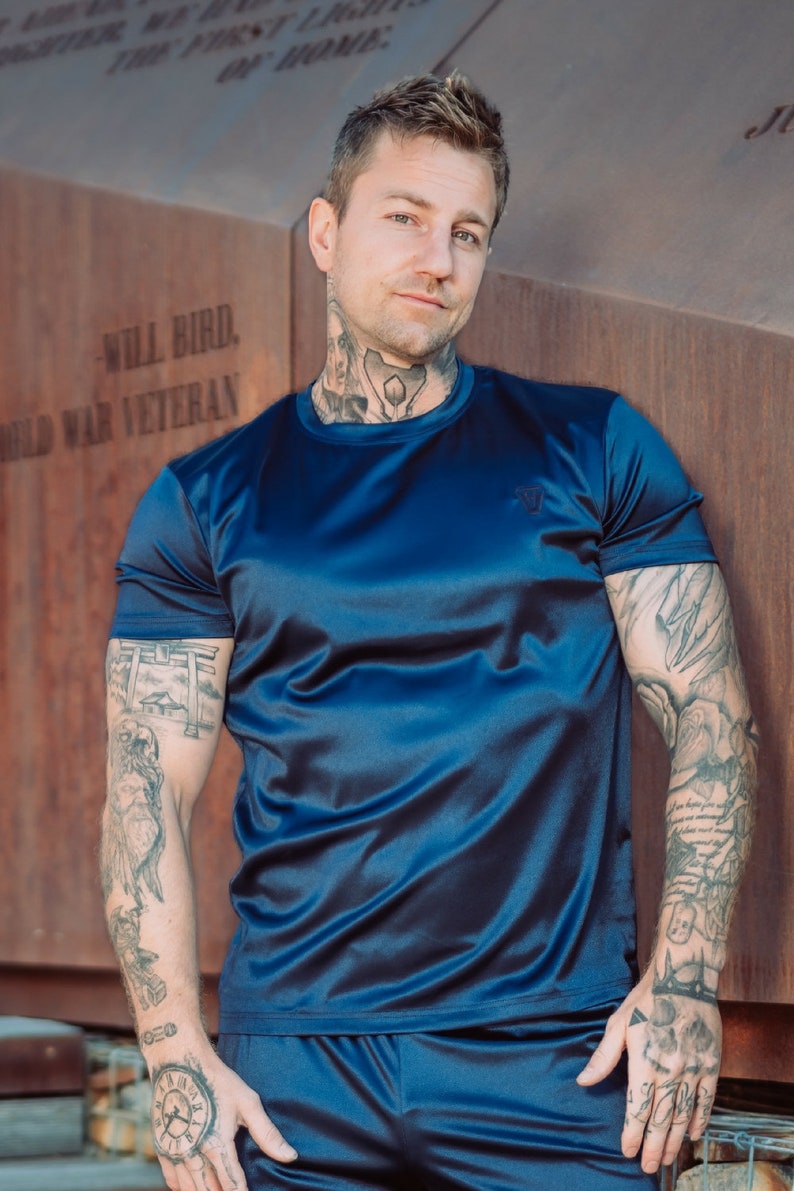 May include: A navy blue athletic t-shirt and shorts set. The short-sleeved shirt has a small logo on the chest. The set is made of a shiny, silky material. The person in the image has tattoos on their arms and neck.