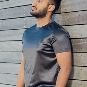 May include: Charcoal gray athletic t-shirt and shorts set. The short-sleeved shirt has a small logo on the chest. The fabric has a subtle sheen. The model is leaning against a wooden wall.