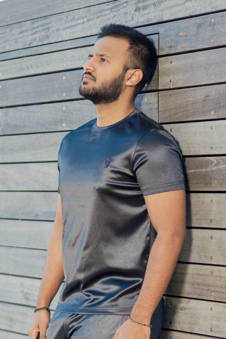 May include: Charcoal gray athletic t-shirt and shorts set. The short-sleeved shirt has a small logo on the chest. The fabric has a subtle sheen. The model is leaning against a wooden wall.