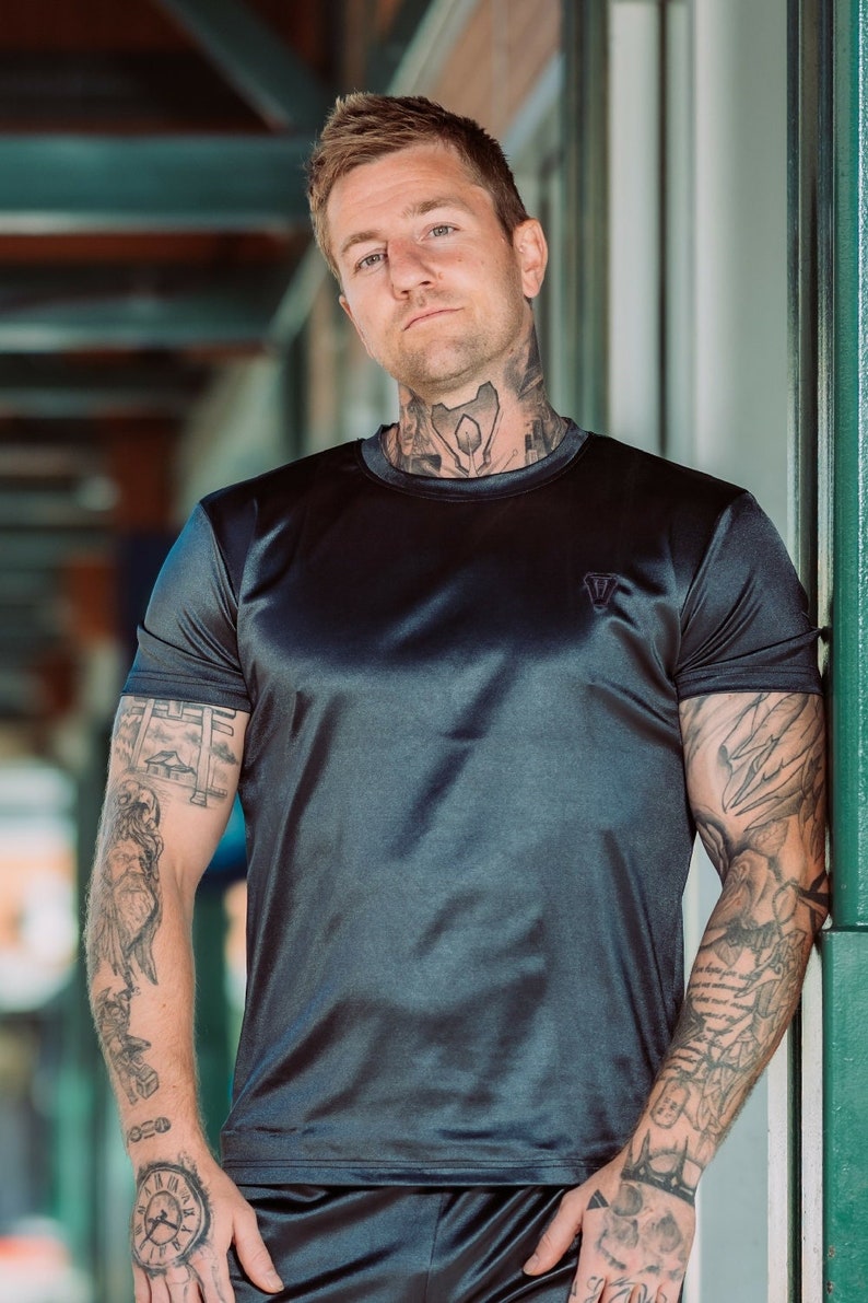May include: A dark gray, short-sleeved t-shirt with a small logo on the chest. The shirt has a silky sheen. The person wearing the shirt has tattoos on their arms and neck. The person is leaning against a green support beam.