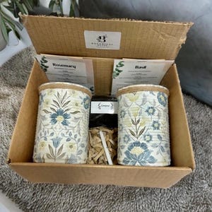 May include: A cardboard gift box containing two cylindrical metal planters with a blue and yellow floral pattern, wrapped with twine at the top. The box includes two instruction cards labelled Rosemary and Basil, a small bag of compost, and a white marker.