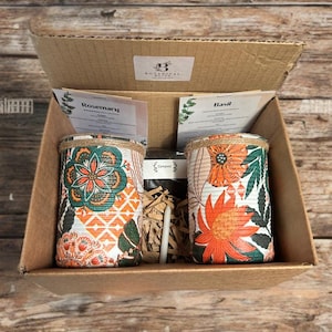 May include: A cardboard box containing two decorative plant pots with floral designs in orange, green, and white. The pots are labelled "Rosemary" and "Basil" and are accompanied by seed packets and compost. The box has a label that says "Botanical Bites".