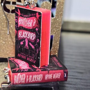 Pode incluir: Um par de brincos com capas de livros em miniatura do romance "A Vida Invis&iacute;vel de Addie LaRue" de V.E. Schwab. As capas dos livros s&atilde;o rosa e preto com o t&iacute;tulo e o nome do autor impressos nelas.