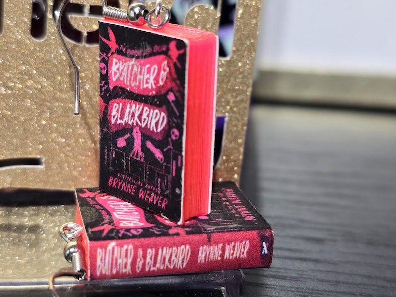 Pode incluir: Um par de brincos com capas de livros em miniatura do romance "A Vida Invis&iacute;vel de Addie LaRue" de V.E. Schwab. As capas dos livros s&atilde;o rosa e preto com o t&iacute;tulo e o nome do autor impressos nelas.