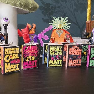 May include: A collection of miniature book charms with vibrant covers, including titles such as "Dungeon Crawler" and "The Butcher's Masquerade." The books are displayed with Lego figures, creating a fantasy theme. The author's name "Matt Dinniman" is visible.