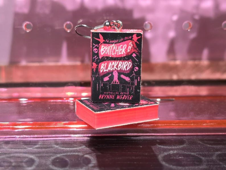Pode incluir: Um par de brincos com capas de livros em miniatura do romance "The Butcher and the Blackbird" de Brynne Weaver. As capas dos livros s&atilde;o rosa e preto com texto branco. Os brincos s&atilde;o prateados.