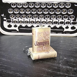 May include: A gold charm with a vintage book cover design. The book cover features the title "Interview with the Vampire" by Anne Rice.
