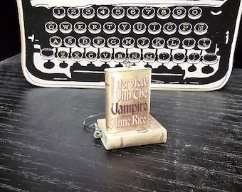 Puede incluir: Un amuleto dorado con un diseño de portada de libro vintage. La portada del libro muestra el título "Entrevista con el vampiro" de Anne Rice.