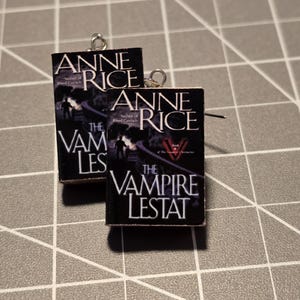 May include: A pair of earrings featuring miniature book covers. The covers display the title "The Vampire Lestat" by Anne Rice, with a dark background and white lettering. The earrings have silver-colored hooks.