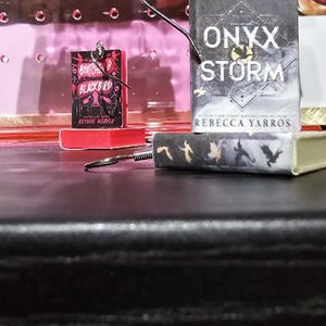 Pode incluir: Dois brincos com tema de livros. O primeiro brinco apresenta um livro em miniatura com o t&iacute;tulo "A  Batched of Blackbird" e o autor "Rebecca Weaver". O segundo brinco apresenta um livro em miniatura com o t&iacute;tulo "Onyx Storm" e o autor "Rebecca Yarros".