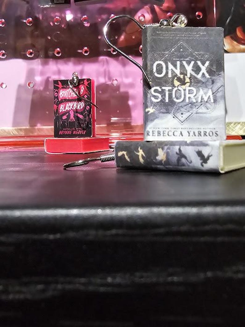 Pode incluir: Dois brincos com tema de livros. O primeiro brinco apresenta um livro em miniatura com o t&iacute;tulo "A  Batched of Blackbird" e o autor "Rebecca Weaver". O segundo brinco apresenta um livro em miniatura com o t&iacute;tulo "Onyx Storm" e o autor "Rebecca Yarros".