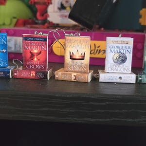 Może przedstawiać: Miniaturowe zawieszki w kształcie książek z okładkami z serii "Gra o Tron" George'a R.R. Martina. Książki mają różne wzory okładek, w tym miecz, kielich, koronę, smoka i burzę. Każda książka jest przymocowana do srebrnej pętelki.