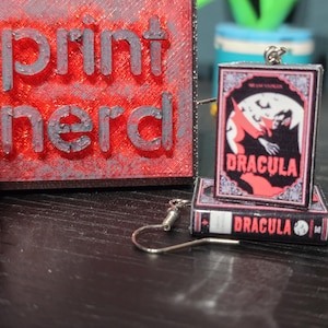 May include: A pair of earrings featuring miniature book charms with the title "Dracula" printed on the front and back. The charms are attached to silver-toned earring hooks.