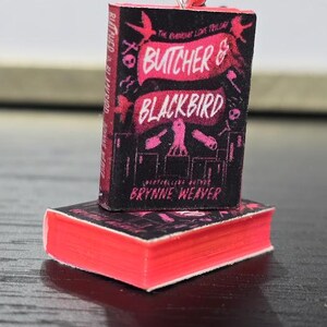 Pode incluir: Um amuleto de livro em miniatura com a capa de "Butcher & Blackbird" de Brynne Weaver. A capa &eacute; preta com texto e ilustra&ccedil;&otilde;es rosa e branco. O amuleto est&aacute; preso a uma corrente prateada.