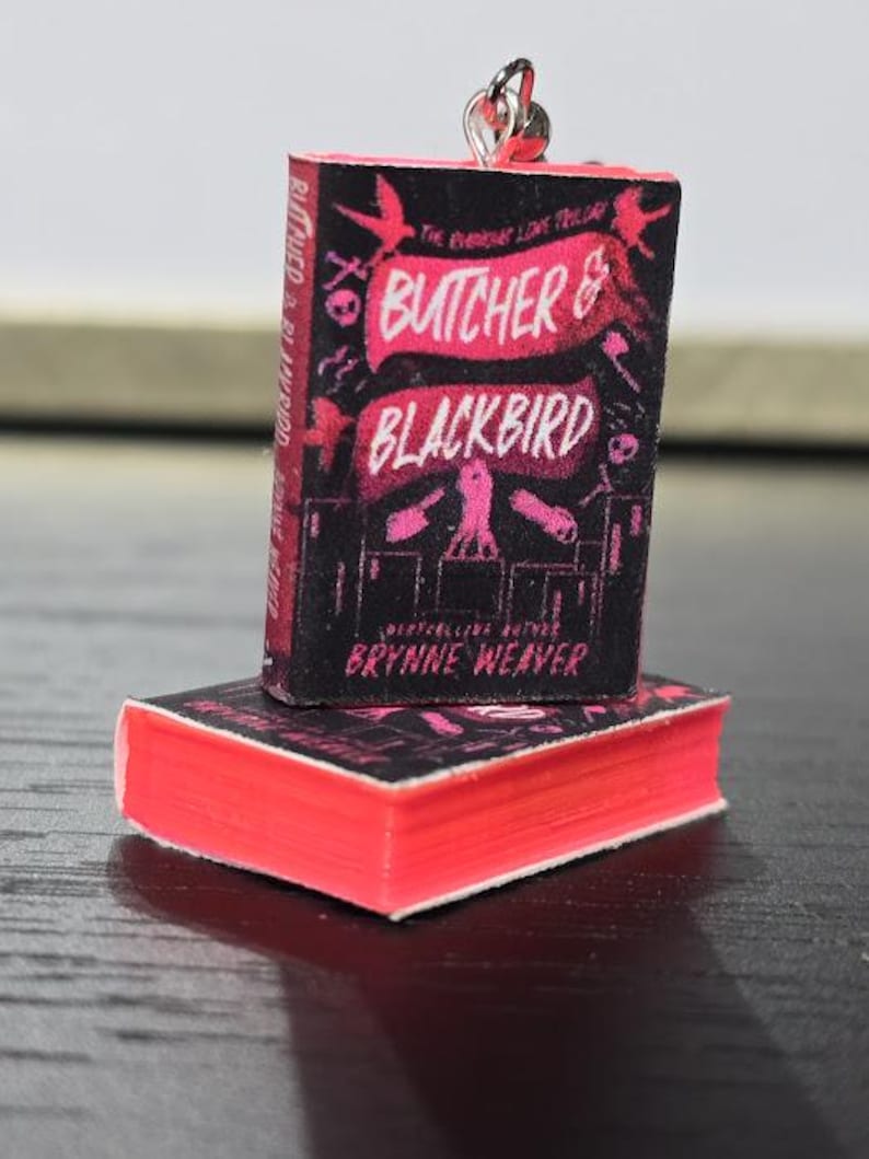 Pode incluir: Um amuleto de livro em miniatura com a capa de "Butcher & Blackbird" de Brynne Weaver. A capa &eacute; preta com texto e ilustra&ccedil;&otilde;es rosa e branco. O amuleto est&aacute; preso a uma corrente prateada.