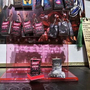 Pode incluir: Um expositor rosa com v&aacute;rios brincos com tema de livros. Os brincos apresentam capas dos livros "Brother Blackbird", "Onyx & Storm" e "Gods of Jade and Shadow". O expositor est&aacute; sobre uma superf&iacute;cie escura com um marcador de p&aacute;gina de madeira que diz "Nossa jornada come&ccedil;ou. Um mago, uma elfa, um an&atilde;o e um guerreiro em busca do reino perdido."