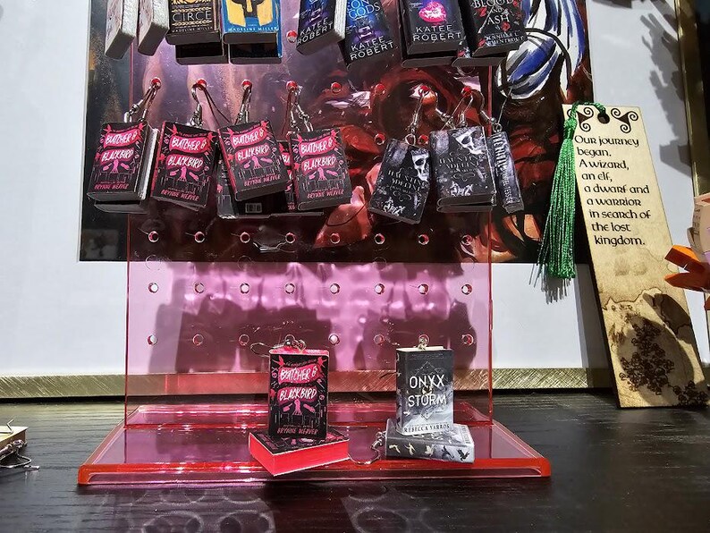 Pode incluir: Um expositor rosa com v&aacute;rios brincos com tema de livros. Os brincos apresentam capas dos livros "Brother Blackbird", "Onyx & Storm" e "Gods of Jade and Shadow". O expositor est&aacute; sobre uma superf&iacute;cie escura com um marcador de p&aacute;gina de madeira que diz "Nossa jornada come&ccedil;ou. Um mago, uma elfa, um an&atilde;o e um guerreiro em busca do reino perdido."