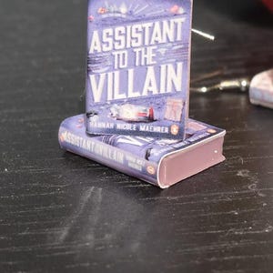 May include: A pair of earrings shaped like miniature books with the title "Assistant to the Villain" by Hannah Nicole  Maenner. The books are purple with a pink spine and are on a dark wood surface.